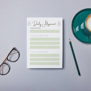 Op de afbeelding: Een wit, printbaar dagelijks uitlijningsblad met een groene achtergrond. Het werkblad heeft een sectie voor de bevestiging van vandaag, drie dingen waar je dankbaar voor bent en drie dingen waar je enthousiast over bent.