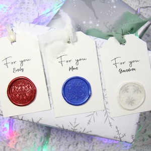 May include: Three white gift tags with handwritten text, "For you" followed by a name, and a wax seal. The seals are red with a Christmas ornament design, blue with a "Merry Christmas" design, and white with a snowflake design.
