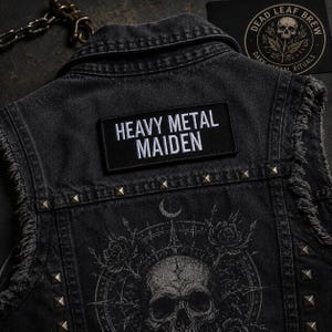 May include: A black denim waistcoat featuring a rectangular patch with the white embroidered text HEAVY METAL MAIDEN. The waistcoat is adorned with silver pyramid studs along the yoke and includes a graphic print of a skull, roses, and a crescent moon.