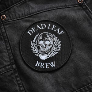 Könnte beinhalten: Ein kreisförmiger, schwarzer Stickaufnäher mit einem weißen Totenkopf in einem Blattkranz und einer kleinen Tasse unter dem Totenkopf. Der Text DEAD LEAF ist über dem Totenkopf gebogen und das Wort BREW steht unter der Tasse auf dem Aufnäher.