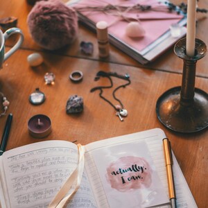 May include: An open journal with a pink and white sticker that says "actually, I can." The journal is on a wooden table with a lit candle, a small bottle, and other decorative items.