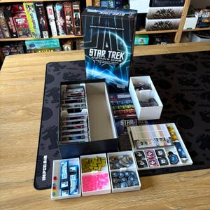 Könnte beinhalten: Ein Star Trek Captain's Chair Brettspiel-Set. Die Box ist schwarz mit blauen und weißen Grafiken. Das Spiel enthält Karten, Spielsteine und Spielteile in verschiedenen Behältern. Das Spiel befindet sich auf einer schwarzen Matte.