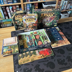 Op de afbeelding: Bordspeldozen en componenten worden getoond op een zwarte mat. De dozen tonen de titel "Arnak" in verschillende ontwerpen. De open doos onthult spelstukken, kaarten en tokens. De achtergrond bevat een boekenplank vol andere bordspellen.