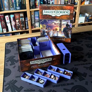 Pode incluir: Uma caixa de jogo de tabuleiro intitulada Arkham Horror Das Kartenspiel Das Fest von Hemlock Vale com um organizador de plástico roxo. O conjunto inclui pilhas de cartas dentro da caixa e três bandejas de plástico roxo separadas com fichas.