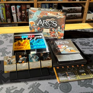 Könnte beinhalten: Ein Brettspiel-Box-Set mit dem Titel "ARCS" mit Weltraum-Thema. Die Box ist geöffnet und zeigt Spielsteine, Karten und ein Regelbuch. Die Karten zeigen Illustrationen und Text. Die Spielsteine befinden sich in gelben und blauen Fächern.