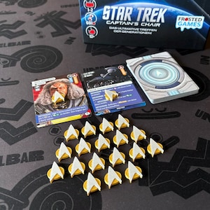 May include: A Star Trek Captain's Chair board game set. The image shows game cards, tokens, and the game box. The cards feature images of characters and game instructions. The tokens are gold and white Starfleet insignia. The box has the game title and logo.
