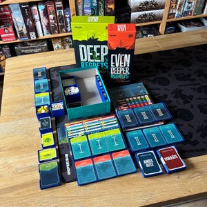 Könnte beinhalten: Ein Brettspiel-Set mit den Schachteln Deep Regrets und Even Deeper Regrets, einer türkisfarbenen Spielschachtel und verschiedenen blauen Kartenhaltern mit Karten, die römische Ziffern, Fischsymbole und deutschen Text aufweisen.