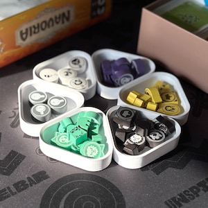 Peut inclure: Un jeu de société avec diverses pièces dans des contenants triangulaires blancs. Les pièces sont de différentes couleurs, dont le blanc, le violet, le jaune, le vert et le noir. La boîte du jeu est visible en arrière-plan, avec le mot "NAUVORIA" imprimé dessus.