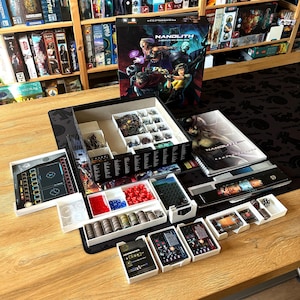 Może przedstawiać: Zestaw gier planszowych, "Nanolith: Cyberpunk RPG Board Game", jest otwarty na drewnianym stole. Grafika na pudełku przedstawia futurystyczne postacie. Zawiera elementy gry, karty, kości i instrukcję. Różne tace zawierają żetony i miniatury.