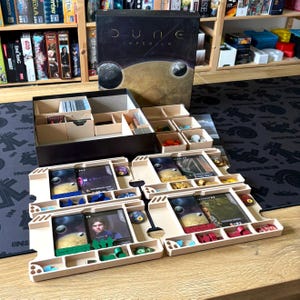 Peut inclure: Organisateur de jeu de société beige pour Dune: Imperium, contenant des cartes, des jetons et d'autres composants. La boîte de jeu est noire avec des lettres dorées et un graphique de planète. D'autres jeux de société sont visibles en arrière-plan.