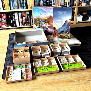 Könnte beinhalten: Ein Brettspiel mit einer Schachtel, die eine Berglandschaft und das Wort "ERDE" zeigt. Das Set enthält Karten, Holzmarker und Tabletts mit Spielsteinen. Die Spielsteine sind in verschiedenen Farben.