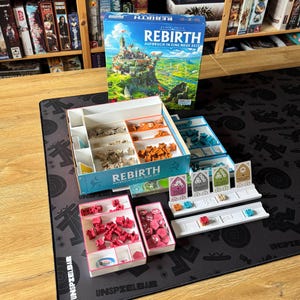 Op de afbeelding: Een bordspel getiteld Rebirth Aufbruch in eine neue Zeit met een speldoos, interne organizers en diverse spelonderdelen, waaronder kleine plastic bouwwerken in beige, oranje, roze en blauw, samen met witte spelersborden en kaarten.