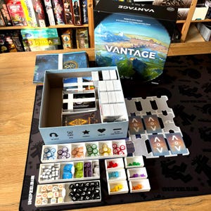 Puede incluir: Juego de mesa "VANTAGE" con una caja que muestra un mapa del mundo. Incluye cartas, fichas y dados, organizados en bandejas. El juego se presenta sobre un tapete de juego negro.