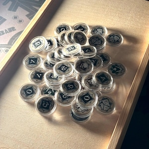 Könnte beinhalten: Eine Ansammlung klarer, runder Spielmarken mit verschiedenen Symbolen und Zahlen, darunter Totenköpfe, Würfel und Sternmuster. Die Spielmarken sind in einer Holzschale angeordnet. Die Zahlen reichen von 0 bis 5.