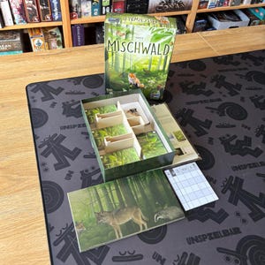 Op de afbeelding: De Mischwald bordspeldoos toont een groene bosillustratie met een vos. De open doos bevat interne verdelers, kaartspellen met bosafbeeldingen, een spelbord met een wolf en das, en een wit scoreblok met een rasterpatroon.