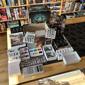 Könnte beinhalten: Ein Brettspiel-Set mit einer großen Schachtel mit Kreaturen-Illustration, zahlreichen weißen Kunststoff-Organizern, grauen Miniaturfiguren, Kartenstapeln, bunten runden Spielsteinen und einem durchsichtigen Kunststoffbehälter mit Farbtöpfen.