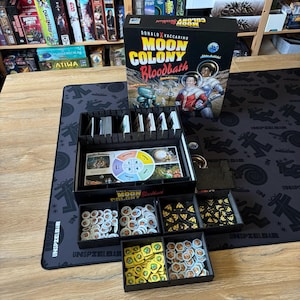 Può includere: Il gioco da tavolo "Moon Colony Bloodbath" con una scatola in bianco e nero. Il gioco è aperto, rivelando pezzi e carte in vassoi neri. Il gioco è su un tappetino nero con un motivo ripetuto.