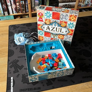 Può includere: Il gioco da tavolo Azul è mostrato, con una scatola decorata con un vivace motivo a piastrelle e il titolo del gioco. All'interno, un vassoio contiene tessere colorate. È presente anche un sacchetto blu con il logo del gioco.