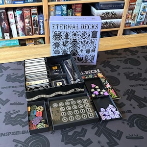 Könnte beinhalten: Brettspiel "Eternal Decks" mit lila-schwarzem Design. Die offene Box zeigt Karten, Spielsteine und Würfel. Das Spielthema ist Fantasy, mit detaillierten Illustrationen auf der Box und den Spielkomponenten.