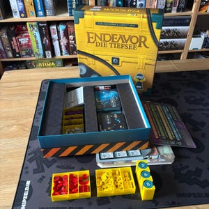 Può includere: Il gioco da tavolo Endeavor Die Tiefsee presenta una scatola gialla con un design a tema sottomarino. La scatola aperta rivela scomparti interni contenenti mazzi di carte, plance giocatore e vassoi in plastica gialla con gettoni rossi e gialli.
