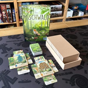 Może przedstawiać: Gra planszowa "MISCHWALD" z pudełkiem przedstawiającym scenę leśną z lisem. Karty do gry z ilustracjami przyrody, talia kart i beżowe pudełko są ułożone na czarnej powierzchni. Widoczny jest tytuł gry.