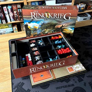 Könnte beinhalten: Das Brettspiel "Der Ringkrieg" wird gezeigt, mit einer Box, die eine Drachenillustration zeigt. Im Inneren sind die Spielkomponenten in Fächern angeordnet: Würfel, Karten und Miniaturfiguren in Rot, Blau und anderen Farben.
