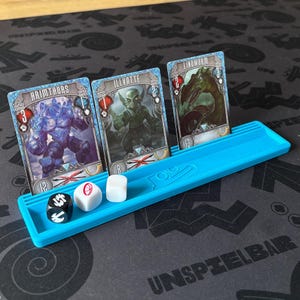 Può includere: Un supporto per carte in plastica blu brillante contenente tre carte da gioco etichettate Hrimthurs, Illvatte e Lindwurm, insieme a tre dadi a sei facce. I dadi includono un dado nero con simboli bianchi e due dadi bianchi con segni rossi e neri.