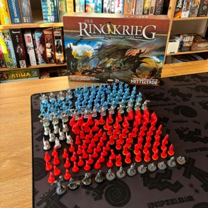 Puede incluir: Una caja de juego de mesa titulada "DER RINGKRIEG" con figuras en miniatura rojas, azules y grises. La caja presenta una ilustración de un dragón. Las piezas del juego están dispuestas sobre una superficie oscura.