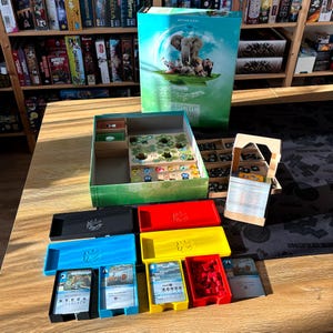 Pode incluir: Uma caixa de jogo de tabuleiro "Arche Nova" com uma ilustração de um elefante e outros animais. O jogo aberto revela peças, cartas e bandejas de várias cores. O jogo está sobre uma mesa de madeira.