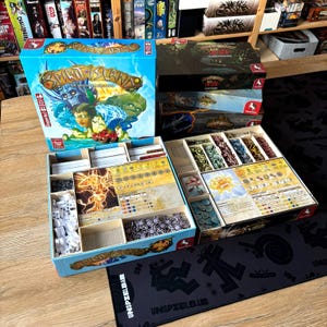 Op de afbeelding: Bordspel "Spirit Island" met een kleurrijke doosillustratie van personages en een landschap. De open doos toont spelcomponenten zoals kaarten en fiches. Andere speldozen zijn ernaast gestapeld. Het spel staat op een houten tafel.