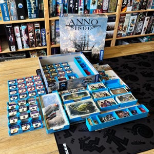 Può includere: Il gioco da tavolo Anno 1800 include una grande scatola illustrata, diversi organizzatori in plastica blu e vari mazzi di carte con ritratti di personaggi e paesaggi. I gettoni e i pezzi di gioco sono ordinati nei vassoi blu.
