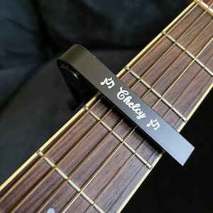 Op de afbeelding: Een zwarte gitaarkapodaster met de tekst "♫ Chelcy ♫" erop gegraveerd. De kapodaster is bevestigd aan het toetsbord van een gitaar.
