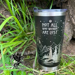 May include: A black stainless steel tumbler with a silver design of mountains, trees, and stars. The tumbler has the text "Not all who wander are lost" on the front.
