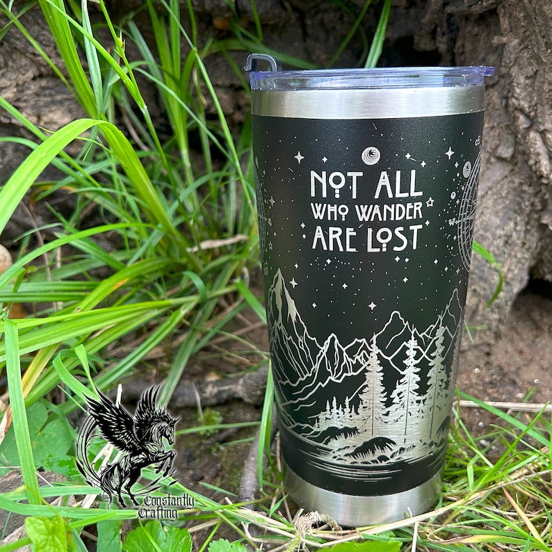 May include: A black stainless steel tumbler with a silver design of mountains, trees, and stars. The tumbler has the text "Not all who wander are lost" on the front.