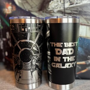 May include: Two stainless steel tumblers with silver lids. One features a detailed black and silver graphic of a spaceship cockpit. The other has the text "THE BEST DAD IN THE GALAXY" in silver lettering on a black background.