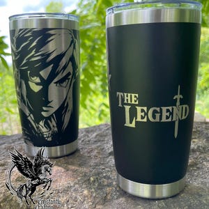 Könnte beinhalten: Zwei Edelstahlbecher mit unterschiedlichen Designs. Ein Becher zeigt eine Schwarzweißillustration einer Figur aus einem Videospiel. Der andere Becher hat einen schwarzen Hintergrund mit dem Text "THE LEGEND" in goldenen Buchstaben und einer Schwertdarstellung.