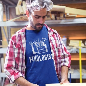 A man wearing safety glasses and a red and white plaid shirt over a royal blue t-shirt with a white graphic of a toolbox and the text "Fixologist".