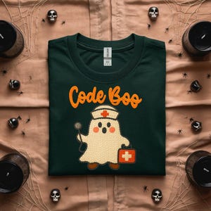 May include: Dark green t-shirt with the text "Code Boo" in orange script. The shirt features a cartoon ghost dressed as a nurse, holding a stethoscope and a medical bag. Halloween-themed decor surrounds the shirt.