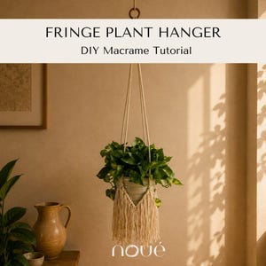 Op de afbeelding: Een crèmekleurige macramé plantenhanger met lange franjes die een groene bladplant vasthoudt. De tekst luidt FRINGE PLANT HANGER DIY Macrame Tutorial en de merknaam nové. De hanger is opgehangen aan een houten ring aan de bovenkant.
