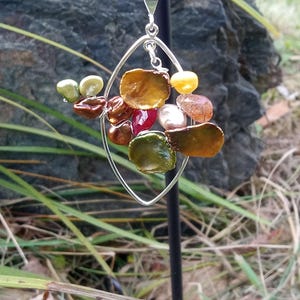 May include: A silver pendant necklace featuring an open oval frame with a cluster of colorful, irregularly shaped beads. The beads are in shades of brown, green, red, yellow, and white. The necklace is displayed outdoors against a natural background.