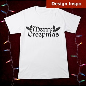 Op de afbeelding: Wit T-shirt met een zwarte afbeelding met de tekst "Merry Creepmas" met schedels en hulstbladeren.
