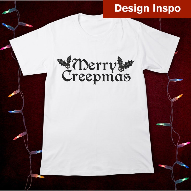 Op de afbeelding: Wit T-shirt met een zwarte afbeelding met de tekst "Merry Creepmas" met schedels en hulstbladeren.