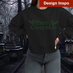 Op de afbeelding: Zwart sweatshirt met een groene "Merry Creepmas" afbeelding met hulst en schedels.
