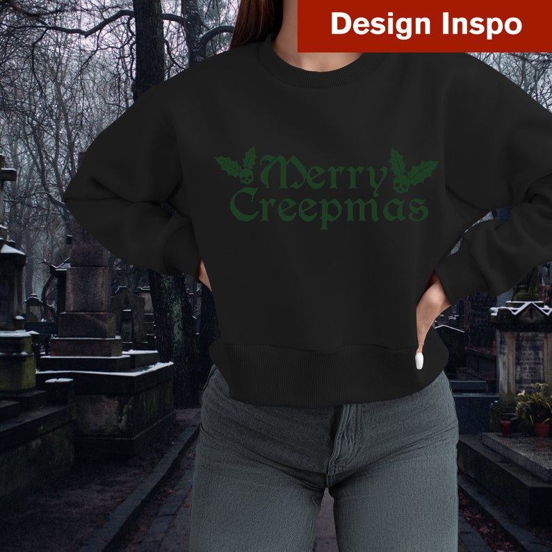 Op de afbeelding: Zwart sweatshirt met een groene "Merry Creepmas" afbeelding met hulst en schedels.