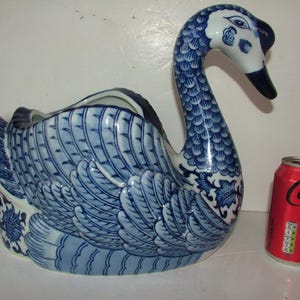 May include: A ceramic swan planter featuring a blue and white hand-painted feather pattern. The swan has a curved neck and a hollow opening on its back. A red Coca-Cola soda can is positioned next to the swan to provide a visual scale reference.
