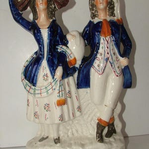 May include: A pair of ceramic figurines depicting a man and a woman in blue and white clothing. The woman is holding a bundle of sticks on her head and the man is holding a stick. The figurines are standing on a white base.