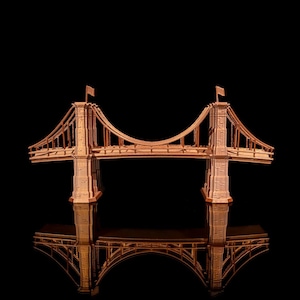 Könnte beinhalten: Ein Holzmodell einer Hängebrücke mit zwei Türmen und einem geschwungenen Deck. Die Brücke spiegelt sich im Wasser darunter.
