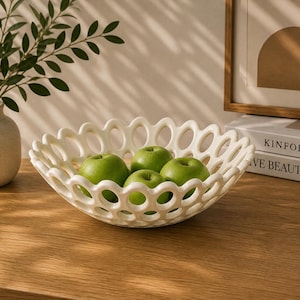 Puede incluir: Un frutero calado de color crema lleno de manzanas verdes se encuentra sobre una superficie de madera. El cuenco tiene un diseño de borde festoneado. Un jarrón con vegetación y una imagen enmarcada están en el fondo.