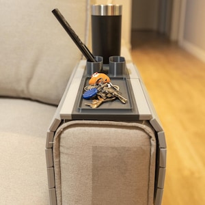 Op de afbeelding: Een grijze armleuning van de bank met ingebouwde bekerhouders en een klein dienblad voor sleutels en andere items. Een zwarte beker, pen en een set sleutels liggen op de lade. De lade is bevestigd aan de arm van een beige bank.