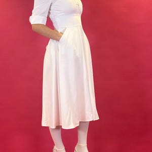 Puede incluir: Un vestido blanco con cuello, mangas cortas y corpiño entallado. El vestido tiene una falda acampanada y bolsillos. La persona lleva medias blancas y tacones de plataforma blancos. El fondo es de color magenta.
